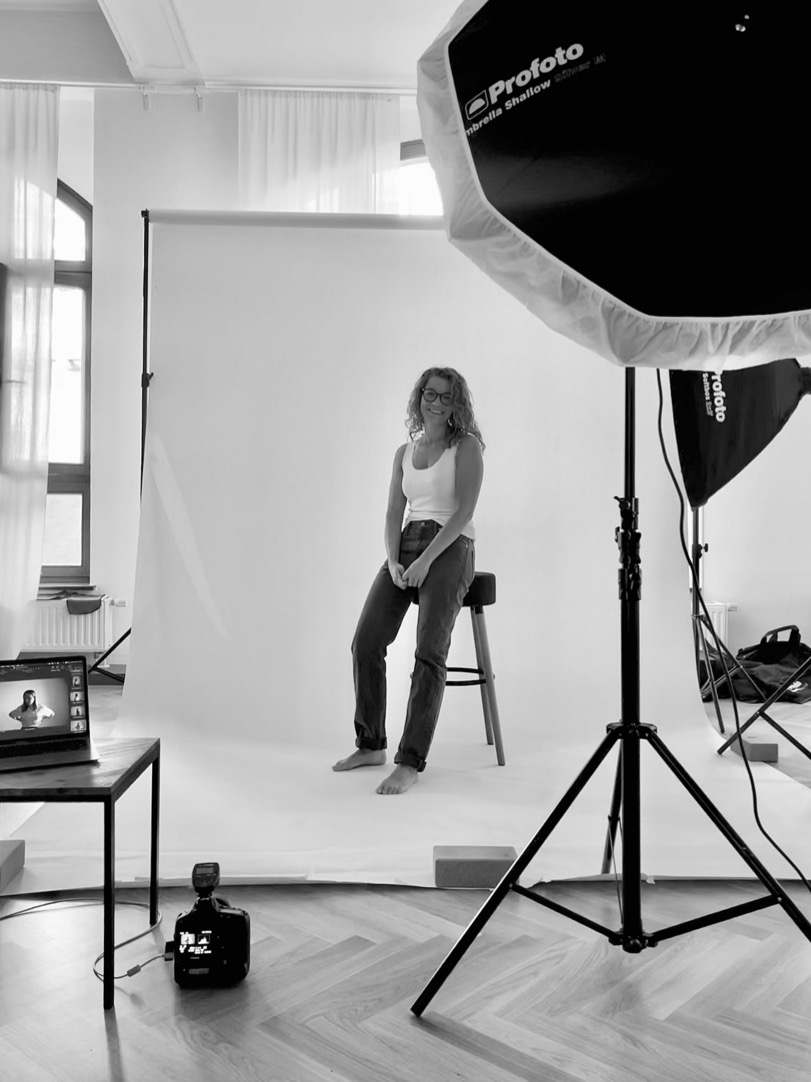 Frau sitzt barfuß auf einem Hocker im Fotostudio vor weißem Hintergrund. Sie trägt Jeans und ein schlichtes Top, wirkt entspannt und natürlich. Umgeben von professionellem Lichtequipment und Kamera – Einblick in die ruhige, authentische Atmosphäre eines Fotoshootings.